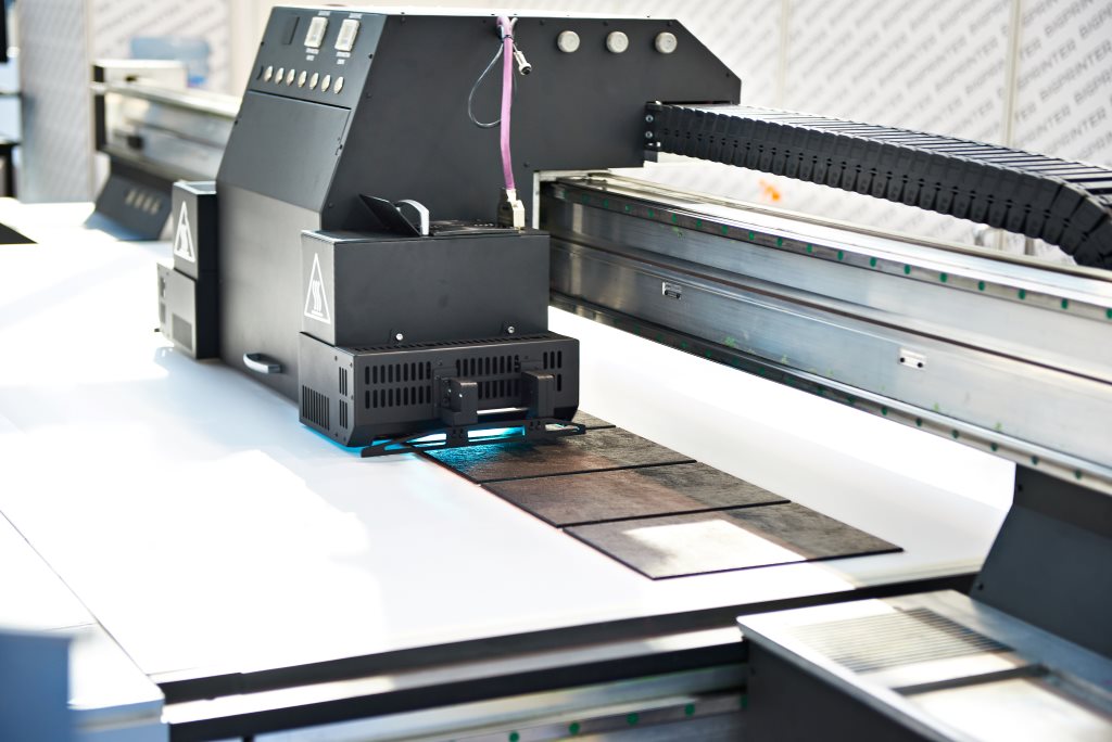 A large-format industrial printer in operation, printing detailed designs onto a flat material surface in a production setting.